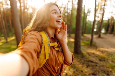 Güneşli bir ormanda selfie çeken sırt çantalı güzel bir kadın yürüyüşçünün portresi. Yürüyüş ve blog yapmaktan hoşlanan genç bir kadın. Macera konsepti. Etkin yaşam biçimi.