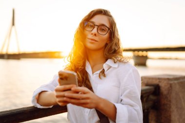 Gün batımında elinde telefonla setin kenarında yürüyen tatlı bir kadın. Dışarıda telefon konuşmaları yapan güzel bir kadın. Blog yazma, boş zaman kavramı.