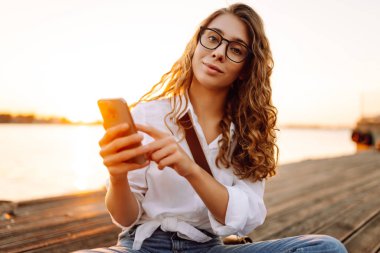 Gün batımında elinde telefonla setin kenarında yürüyen tatlı bir kadın. Dışarıda telefon konuşmaları yapan güzel bir kadın. Blog yazma, boş zaman kavramı.