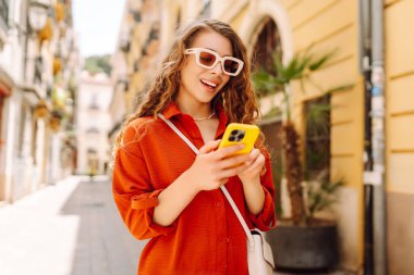 Güneş gözlüklü, elinde telefonla güneşli caddelerde yürüyen güzel bir kadının portresi. Turist dışarıda havanın tadını çıkarıyor. Seyahat kavramı.