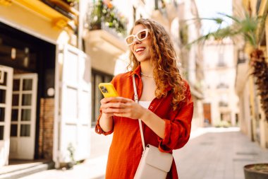 Güneş gözlüklü, elinde telefonla güneşli caddelerde yürüyen güzel bir kadının portresi. Turist dışarıda havanın tadını çıkarıyor. Seyahat kavramı.