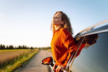 Genç kadın arabanın camından sarkan özgürlüğü hissediyor. Güzel bayan gezgin arabadan gün batımının tadını çıkarıyor. Araba yolculuğu, eğlence konsepti. Etkin yaşam biçimi.