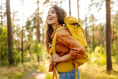 Sarı sırt çantalı kadın yürüyüşçü güneşli ormanda yürürken günışığının tadını çıkarıyor. Yürüyüş yolunda genç bir kadın yürüyüşçü. Turizm, yürüyüş konsepti.