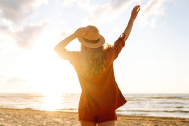 Sahilde gün batımında eğlenen şapkalı genç bir turist. Özgürlüğü hisseden ve deniz manzarasının tadını çıkaran güzel bir kadın. Yürüyüş, dinlenme konsepti.