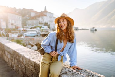 Şapkalı neşeli turist setin üzerinde güneşin doğuşunun tadını çıkarıyor. Genç bir kadın güzel bir mimarisi olan şirin bir köyde yürüyor. Turizm, seyahat konsepti. Etkin yaşam biçimi.