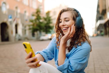Kablosuz kulaklıklı ve elinde telefon olan mutlu kadın güneşli Avrupa caddelerinde yürüyor. Genç turist dışarıda eğleniyor. Yürüme, dinlenme konsepti. Etkin yaşam biçimi.