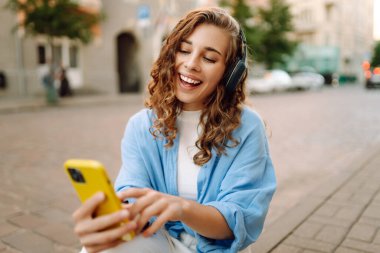 Kablosuz kulaklıklı ve elinde telefon olan mutlu kadın güneşli Avrupa caddelerinde yürüyor. Genç turist dışarıda eğleniyor. Yürüme, dinlenme konsepti. Etkin yaşam biçimi.