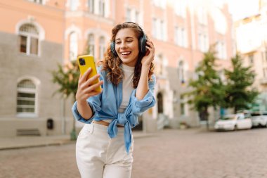 Kablosuz kulaklıklı ve elinde telefon olan mutlu kadın güneşli Avrupa caddelerinde yürüyor. Genç turist dışarıda eğleniyor. Yürüme, dinlenme konsepti. Etkin yaşam biçimi.