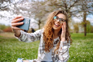 Selfie zamanı. Güzel bir kadın çiçek açan bir bahçede modern bir telefonla fotoğraf çekiyor. Kadın güneşli bir bahar gününün tadını çıkarıyor. Teknoloji kavramı, yürüyüşler, rahatlama..