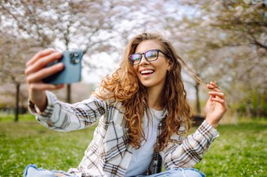 Selfie zamanı. Güzel bir kadın çiçek açan bir bahçede modern bir telefonla fotoğraf çekiyor. Kadın güneşli bir bahar gününün tadını çıkarıyor. Teknoloji kavramı, yürüyüşler, rahatlama..