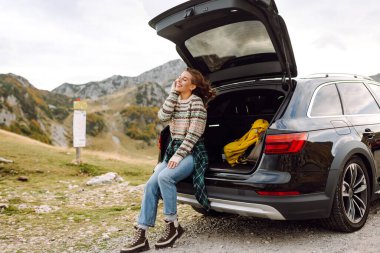 Gün batımında bir arabanın bagajında dağ manzarasının tadını çıkaran güzel bir kadın. Genç bayan bir gezgin bir arabanın bagajında özgürce dolaşıyor. Seyahat tutkusu.