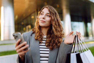 Alışveriş merkezinin yanındaki telefon ve alışveriş torbalarıyla güzel bir kadının portresi. Şık bir kadın internette telefon alıp açık havada günbatımının tadını çıkarıyor. Çevrimiçi alışveriş, satın alma kavramı.