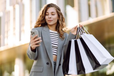 Alışveriş merkezinin yanındaki telefon ve alışveriş torbalarıyla güzel bir kadının portresi. Şık bir kadın internette telefon alıp açık havada günbatımının tadını çıkarıyor. Çevrimiçi alışveriş, satın alma kavramı.