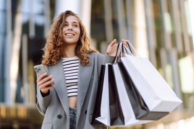 Alışveriş merkezinin yanındaki telefon ve alışveriş torbalarıyla güzel bir kadının portresi. Şık bir kadın internette telefon alıp açık havada günbatımının tadını çıkarıyor. Çevrimiçi alışveriş, satın alma kavramı.