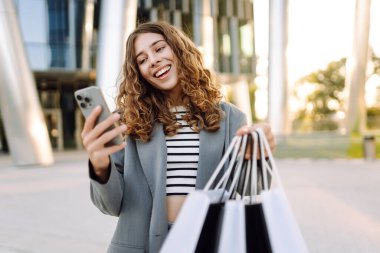 Alışveriş merkezinin yanındaki telefon ve alışveriş torbalarıyla güzel bir kadının portresi. Şık bir kadın internette telefon alıp açık havada günbatımının tadını çıkarıyor. Çevrimiçi alışveriş, satın alma kavramı.