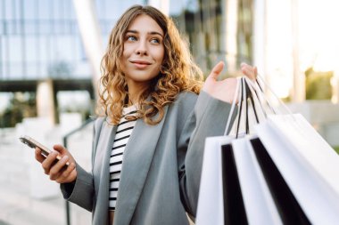 Alışveriş merkezinin yanındaki telefon ve alışveriş torbalarıyla güzel bir kadının portresi. Şık bir kadın internette telefon alıp açık havada günbatımının tadını çıkarıyor. Çevrimiçi alışveriş, satın alma kavramı.
