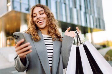 Alışveriş merkezinin yanındaki telefon ve alışveriş torbalarıyla güzel bir kadının portresi. Şık bir kadın internette telefon alıp açık havada günbatımının tadını çıkarıyor. Çevrimiçi alışveriş, satın alma kavramı.