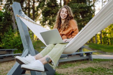 Dizüstü bilgisayarı olan genç bir kadın temiz havada ağaçların arasında bir hamakta yatıyor. Serbest çalışan bir kadın gün batımında dizüstü bilgisayarla parkta vakit geçiriyor. Serbest çalışma, özgürlük kavramı. Etkin yaşam biçimi.