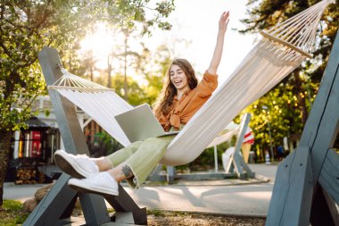 Dizüstü bilgisayarı olan genç bir kadın temiz havada ağaçların arasında bir hamakta yatıyor. Serbest çalışan bir kadın gün batımında dizüstü bilgisayarla parkta vakit geçiriyor. Serbest çalışma, özgürlük kavramı. Etkin yaşam biçimi.