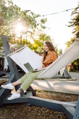 Dizüstü bilgisayarı olan genç bir kadın temiz havada ağaçların arasında bir hamakta yatıyor. Serbest çalışan bir kadın gün batımında dizüstü bilgisayarla parkta vakit geçiriyor. Serbest çalışma, özgürlük kavramı. Etkin yaşam biçimi.