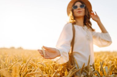 Gün batımı tarlasında altın buğdaya dokunan genç bir kadının elleri. Tarlada gezen şık elbiseli güzel bir kadın. Yürüme ve eğlenme kavramı.