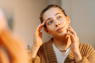 Yüzüne nemlendirici süren genç bir kadının portresi. Güzel bir kadın aynadaki kusurlarını inceliyor. Güzellik ve doğallık kavramı.