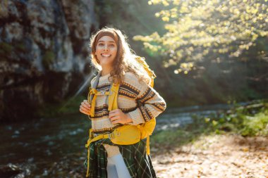 Dağlarda dağ nehrinin yanında yürüyüş yapan sarı sırt çantalı genç bir kadın. Mutlu bayan gezgin açık havada güneşli havanın tadını çıkarıyor. Seyahat konsepti, yürüyüş. Etkin yaşam biçimi.