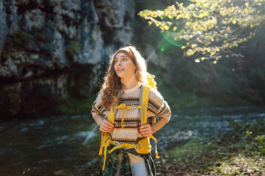Dağlarda dağ nehrinin yanında yürüyüş yapan sarı sırt çantalı genç bir kadın. Mutlu bayan gezgin açık havada güneşli havanın tadını çıkarıyor. Seyahat konsepti, yürüyüş. Etkin yaşam biçimi.