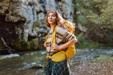 Dağlarda dağ nehrinin yanında yürüyüş yapan sarı sırt çantalı genç bir kadın. Mutlu bayan gezgin açık havada güneşli havanın tadını çıkarıyor. Seyahat konsepti, yürüyüş. Etkin yaşam biçimi.