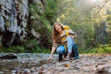 Dağlarda dağ nehrinin yanında yürüyüş yapan sarı sırt çantalı genç bir kadın. Mutlu bayan gezgin açık havada güneşli havanın tadını çıkarıyor. Seyahat konsepti, yürüyüş. Etkin yaşam biçimi.