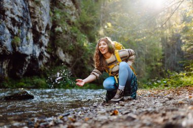 Dağlarda dağ nehrinin yanında yürüyüş yapan sarı sırt çantalı genç bir kadın. Mutlu bayan gezgin açık havada güneşli havanın tadını çıkarıyor. Seyahat konsepti, yürüyüş. Etkin yaşam biçimi.