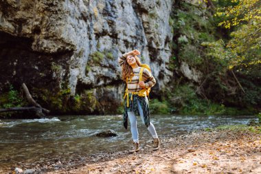 Dağlarda dağ nehrinin yanında yürüyüş yapan sarı sırt çantalı genç bir kadın. Mutlu bayan gezgin açık havada güneşli havanın tadını çıkarıyor. Seyahat konsepti, yürüyüş. Etkin yaşam biçimi.