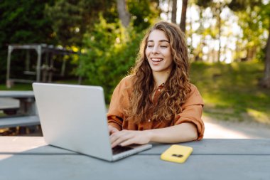 Mutlu kadın parkta dizüstü bilgisayarla oturuyor. Açık havada çalışan serbest çalışan genç bir kadının portresi. Çevrimiçi, teknoloji kavramı