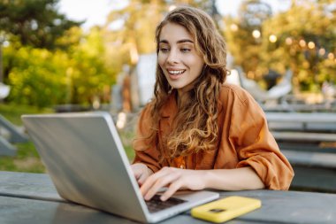 Mutlu kadın parkta dizüstü bilgisayarla oturuyor. Açık havada çalışan serbest çalışan genç bir kadının portresi. Çevrimiçi, teknoloji kavramı