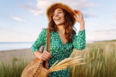 Kumsalda uzun otların yanında yürüyen mutlu bir kadın. Uçan saçlar. Açık havada. Moda tarzı