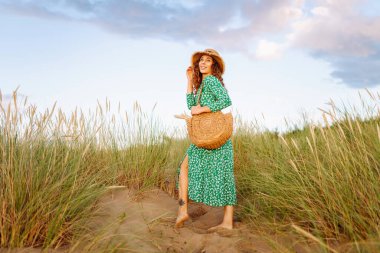 Kumsalda uzun otların yanında yürüyen mutlu bir kadın. Uçan saçlar. Açık havada. Moda tarzı