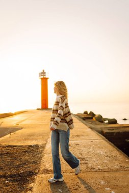 Genç bir kadın gün batımında canlı kırmızı bir deniz fenerinin yanında kumsalda esintili bir günün tadını çıkarıyor. Seyahat, hafta sonu, rahatlama ve yaşam tarzı konsepti