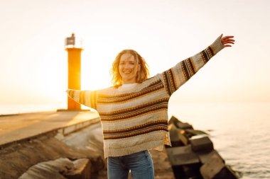 Genç bir kadın gün batımında canlı kırmızı bir deniz fenerinin yanında kumsalda esintili bir günün tadını çıkarıyor. Seyahat, hafta sonu, rahatlama ve yaşam tarzı konsepti
