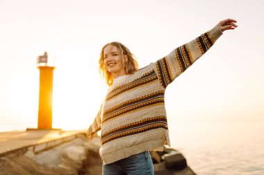 Genç bir kadın gün batımında canlı kırmızı bir deniz fenerinin yanında kumsalda esintili bir günün tadını çıkarıyor. Seyahat, hafta sonu, rahatlama ve yaşam tarzı konsepti