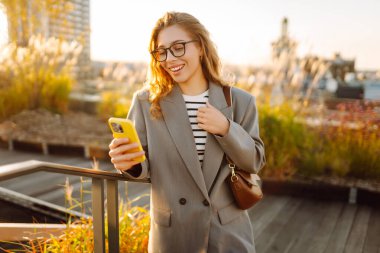 Şehirde gülümseyen ve cep telefonu kullanan mutlu öğrenci kadın. Yaşam tarzı, seyahat, turizm, teknoloji, blog yazma, iletişim kavramı
