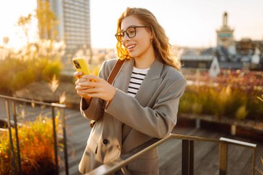 Şehirde gülümseyen ve cep telefonu kullanan mutlu öğrenci kadın. Yaşam tarzı, seyahat, turizm, teknoloji, blog yazma, iletişim kavramı