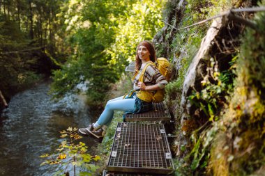 Kadın turist yazın doğayı keşfediyor, köprüde yürüyor. Ormanlarda dolaşan büyük sırt çantalı bir yürüyüşçü. Etkin yaşam biçimi
