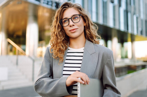 Улыбающаяся женщина, идущая по городской улице с ноутбуком, идет на работу утром. Бизнес, образование, фрилансер