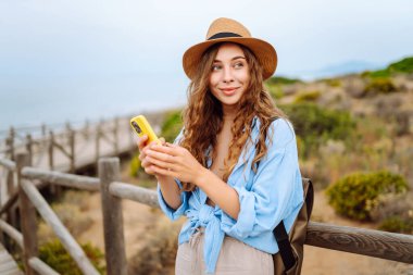 Mutlu kadın akıllı telefon kullanıyor ve deniz kenarında güneşli bir gezinti güvertesinde gülümsüyor. Boş zaman, açık hava ve doğaya yakın. Avrupa seyahati
