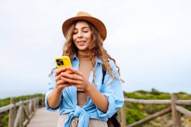 Mutlu kadın akıllı telefon kullanıyor ve deniz kenarında güneşli bir gezinti güvertesinde gülümsüyor. Boş zaman, açık hava ve doğaya yakın. Avrupa seyahati