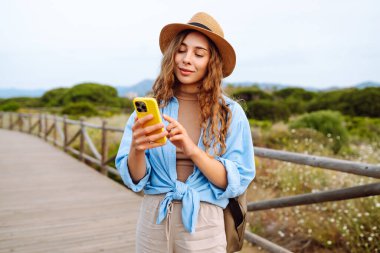 Mutlu kadın akıllı telefon kullanıyor ve deniz kenarında güneşli bir gezinti güvertesinde gülümsüyor. Boş zaman, açık hava ve doğaya yakın. Avrupa seyahati