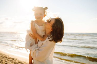 Gülümseyen anne ve güzel kız gün batımında kumsalda eğleniyorlar. Çocukluk, eğlence, rahatlama konsepti