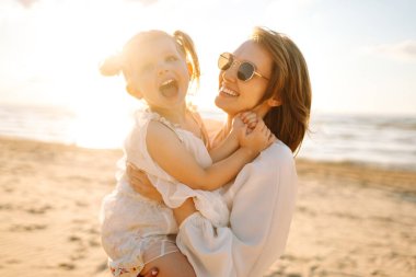 Gülümseyen anne ve güzel kız gün batımında kumsalda eğleniyorlar. Çocukluk, eğlence, rahatlama konsepti