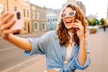 Şık gözlüklü ve modern elbiseli mutlu bir kadın güneşli bir sokakta selfie çekiyor. Yürüme, eğlence, blog yazma, seyahat kavramı