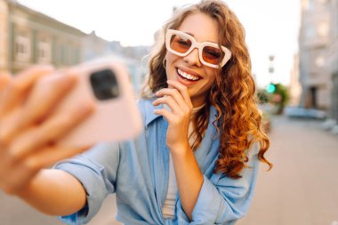 Şık gözlüklü ve modern elbiseli mutlu bir kadın güneşli bir sokakta selfie çekiyor. Yürüme, eğlence, blog yazma, seyahat kavramı